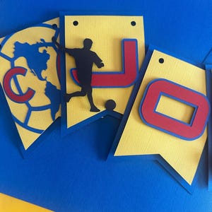 May include: A yellow and blue paper banner with the letters "JO" in red and blue. A silhouette of a soccer player is on the left side of the banner.