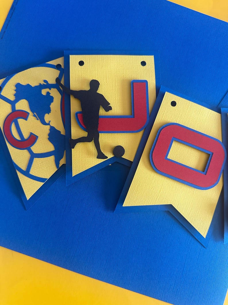 May include: A yellow and blue paper banner with the letters "JO" in red and blue. A silhouette of a soccer player is on the left side of the banner.