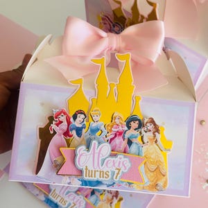 May include: A pink and white favor box with a yellow castle cutout and a pink bow. The box features a scene of Disney princesses with the text "Alexis turns 7".