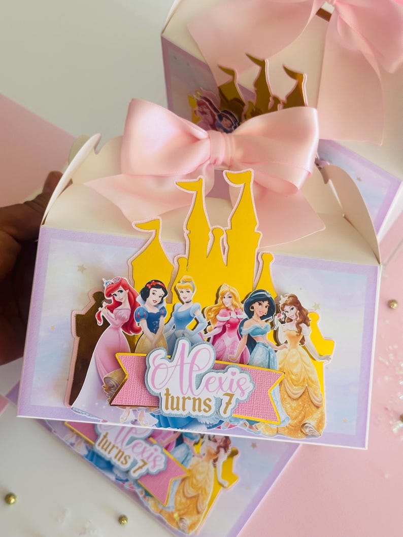 May include: A pink and white favor box with a yellow castle cutout and a pink bow. The box features a scene of Disney princesses with the text "Alexis turns 7".