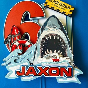 May include: A layered paper cake topper featuring a large red number 6, a shark with an open mouth, a swimmer, a red life buoy, a red rescue float, and a yellow sign reading BEACH CLOSED BY ORDER OF POLICE. The name JAXON is printed in red letters.