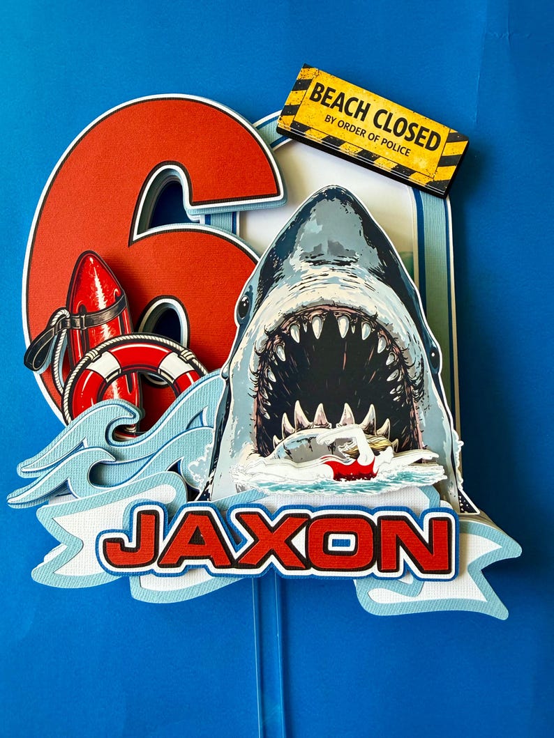 May include: A layered paper cake topper featuring a large red number 6, a shark with an open mouth, a swimmer, a red life buoy, a red rescue float, and a yellow sign reading BEACH CLOSED BY ORDER OF POLICE. The name JAXON is printed in red letters.