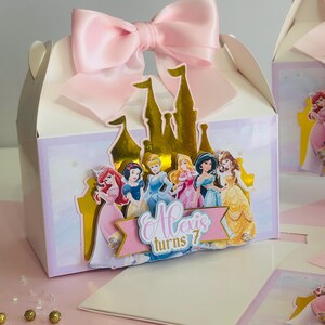 May include: A white favor box with a pink satin bow on top. The box features a gold castle with Disney Princesses on it. The text "Alexis turns 7" is on a pink banner.
