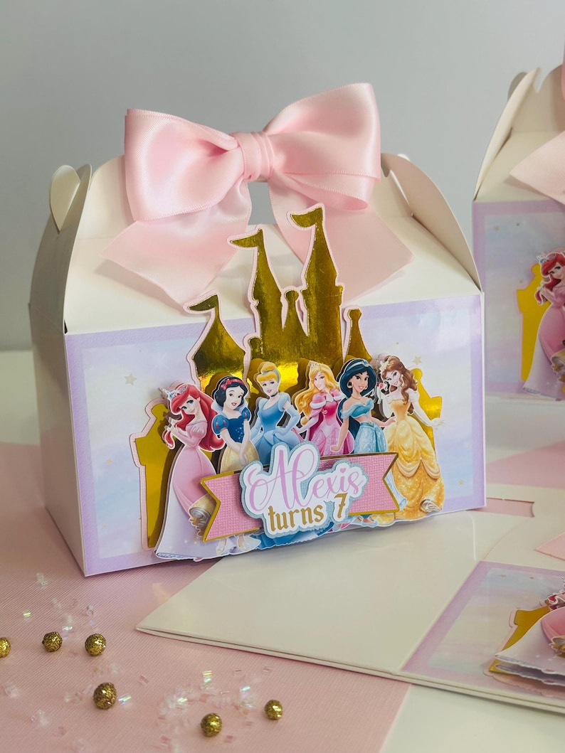 May include: A white favor box with a pink satin bow on top. The box features a gold castle with Disney Princesses on it. The text "Alexis turns 7" is on a pink banner.