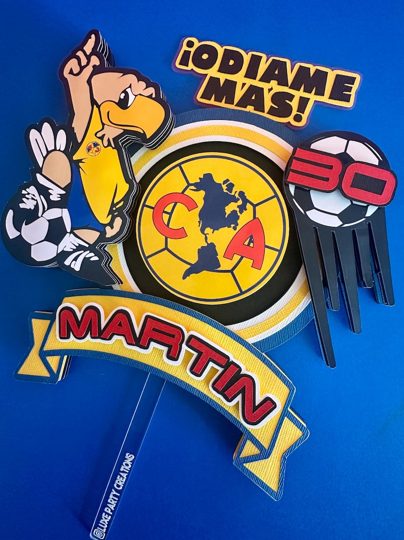 K&ouml;nnte beinhalten: Ein gelber und blauer Tortendekor mit einem Fu&szlig;ball, einem Cartoon-Vogel und den Worten "&iexcl;DIAME MAS!" und "MARTIN".
