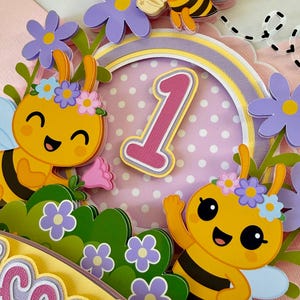 May include: A colorful, layered papercraft cake topper featuring cartoon bees, flowers, and the number 1. The bees are yellow and black with blue wings, and the flowers are purple and pink. The background is pink with white polka dots.