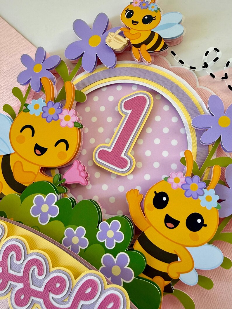 May include: A colorful, layered papercraft cake topper featuring cartoon bees, flowers, and the number 1. The bees are yellow and black with blue wings, and the flowers are purple and pink. The background is pink with white polka dots.