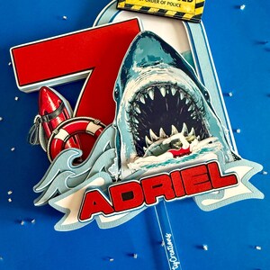 May include: A layered cake topper featuring a large red number 7, a shark with an open mouth, a red and white life buoy, and a yellow sign reading BEACH CLOSED BY ORDER OF POLICE. The name ADRIEL is displayed in red letters on a white banner.