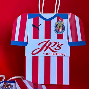 May include: A red and white striped gift bag shaped like a soccer jersey, with a blue collar and sleeve cuffs. The bag features the text "JR's 13th Birthday" and a team logo. White rope handles are attached.