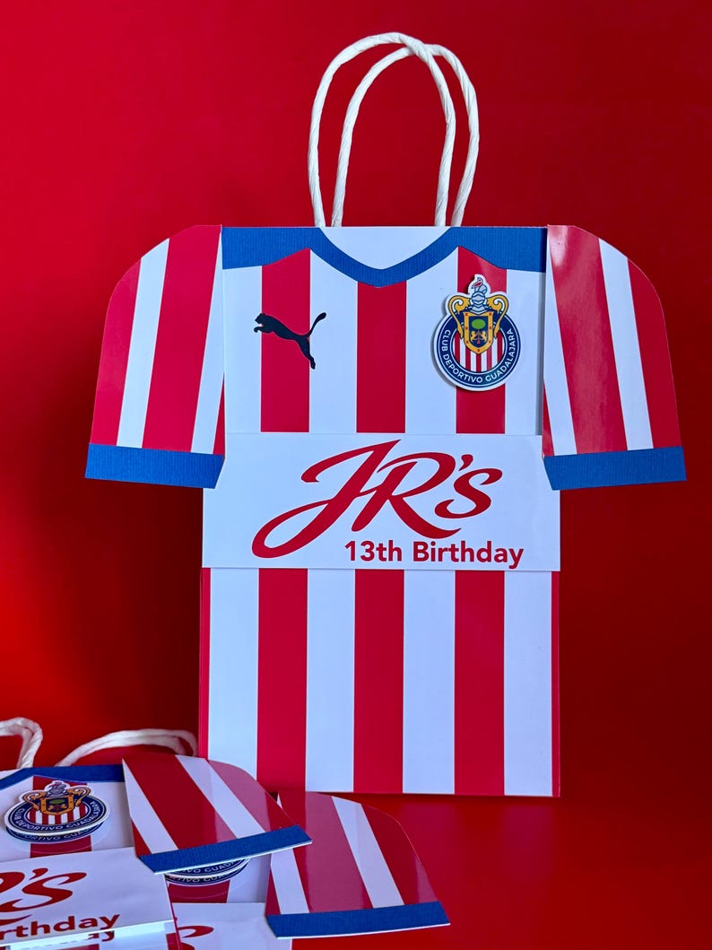 May include: A red and white striped gift bag shaped like a soccer jersey, with a blue collar and sleeve cuffs. The bag features the text "JR's 13th Birthday" and a team logo. White rope handles are attached.