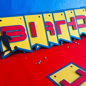 May include: A yellow and blue birthday banner with red letters spelling out "BIRTHDAY". A silhouette of a soccer player is on the left side of the banner.