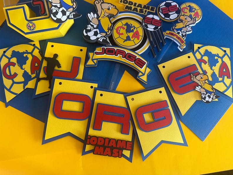 May include: A yellow and blue banner with the letters "JORGE" in red and the words "&iexcl;DI&Aacute;ME M&Aacute;S!" in red. The banner is decorated with soccer balls, a soccer player, and a cartoon bird.