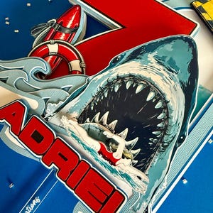 May include: A layered paper cake topper featuring a large shark with an open mouth, a swimmer in a red swimsuit, a red surfboard, a life preserver, and blue waves. The name ADRIEL is printed in bold red letters with white outlines.