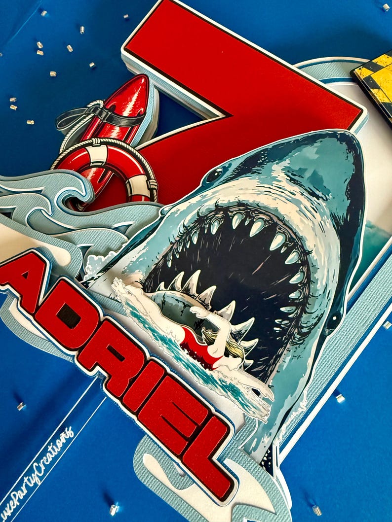 May include: A layered paper cake topper featuring a large shark with an open mouth, a swimmer in a red swimsuit, a red surfboard, a life preserver, and blue waves. The name ADRIEL is printed in bold red letters with white outlines.