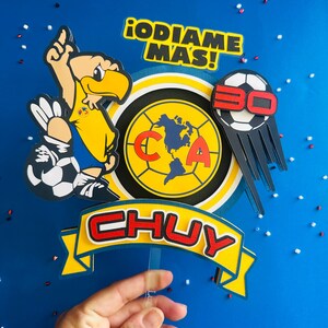 K&ouml;nnte beinhalten: Ein gelber, blauer und roter Tortenaufsatz mit einem Cartoon-Adler, der einen Fu&szlig;ball h&auml;lt, einem Fu&szlig;ball mit der Nummer 30 darauf, einem kreisf&ouml;rmigen Logo mit den Buchstaben "CA" und einer Weltkarte darin sowie einem Banner mit dem Namen "CHUY" in roten Buchstaben. Der Text "&iexcl;ODIAME MAS!" ist ebenfalls auf dem Aufsatz enthalten.