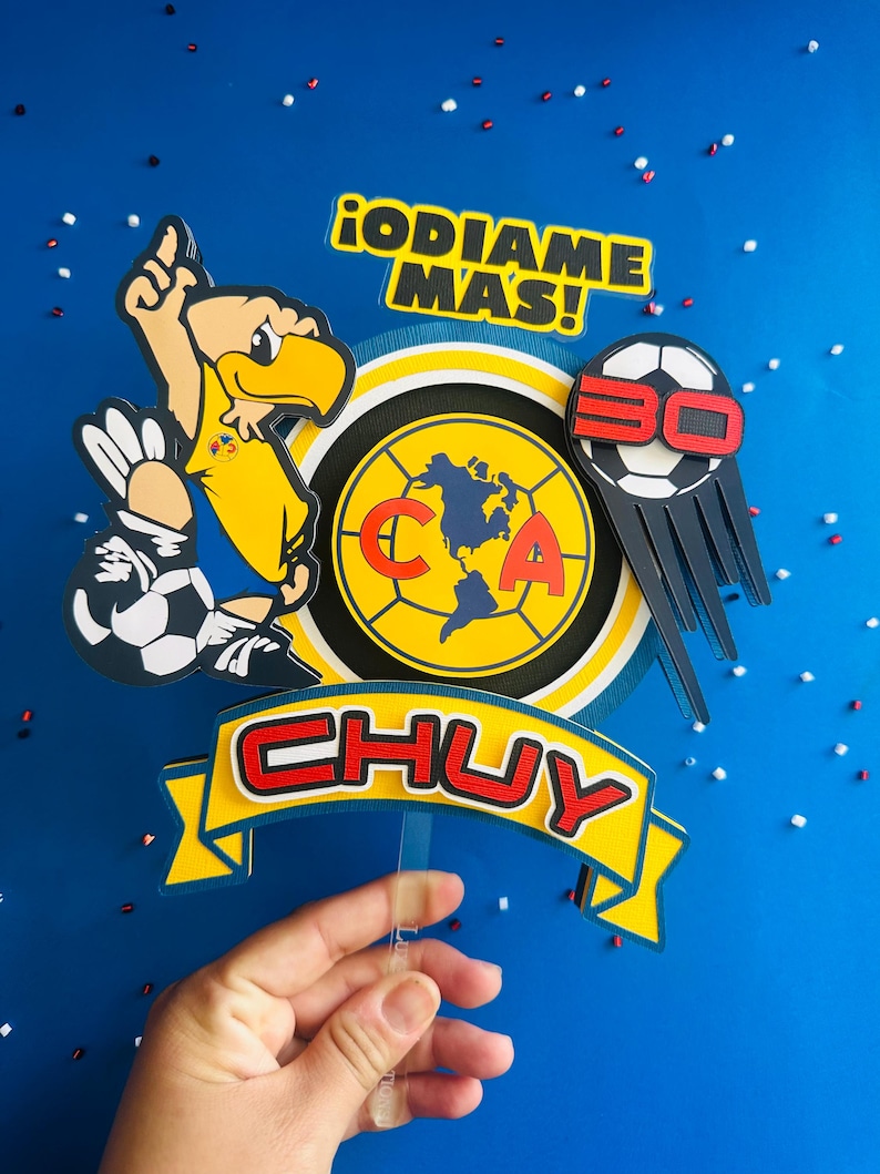 K&ouml;nnte beinhalten: Ein gelber, blauer und roter Tortenaufsatz mit einem Cartoon-Adler, der einen Fu&szlig;ball h&auml;lt, einem Fu&szlig;ball mit der Nummer 30 darauf, einem kreisf&ouml;rmigen Logo mit den Buchstaben "CA" und einer Weltkarte darin sowie einem Banner mit dem Namen "CHUY" in roten Buchstaben. Der Text "&iexcl;ODIAME MAS!" ist ebenfalls auf dem Aufsatz enthalten.