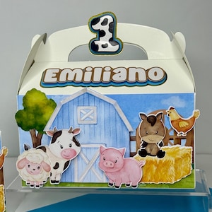 May include: A white party favor box with a farm theme. The box features illustrations of a cow, sheep, pig, and horse in front of a blue barn. The name "Emiliano" is printed on the box, along with a number "1" on top.