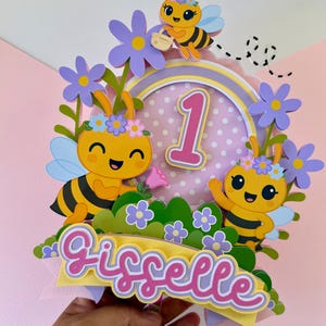 May include: A colorful birthday cake topper featuring cartoon bees, flowers, and the number 1. The topper includes the name "Gisselle" in a cursive font, with a pink and yellow color scheme. The bees have black stripes and are decorated with flowers.