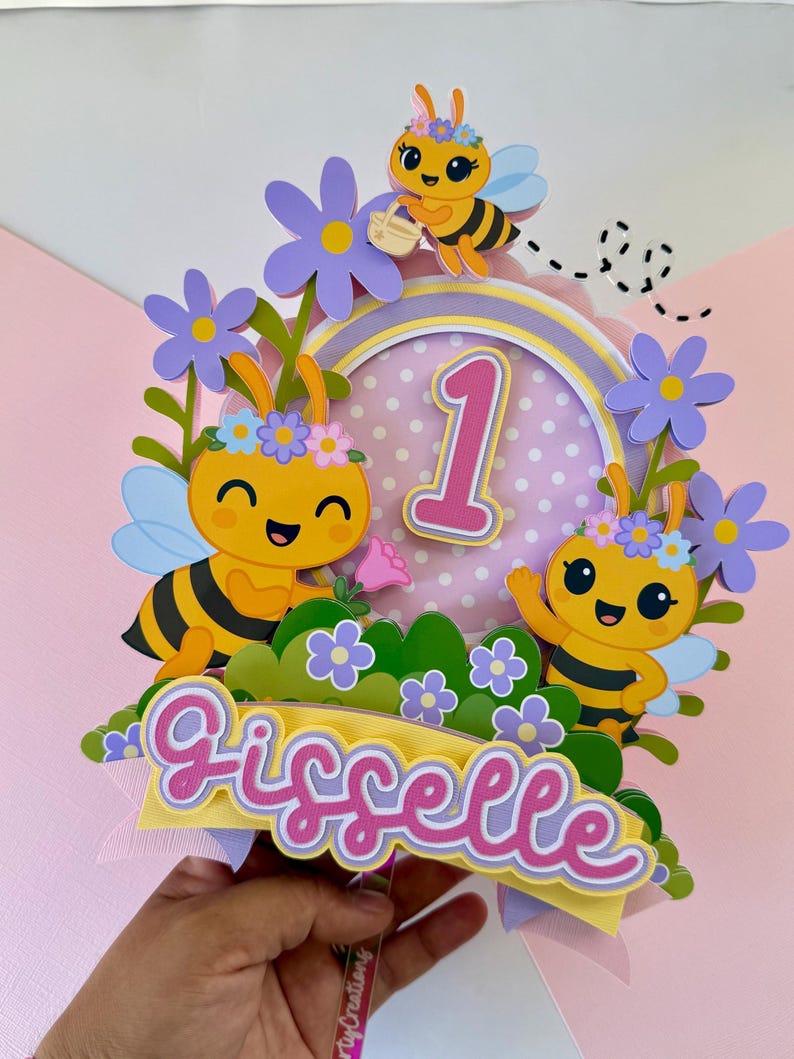May include: A colorful birthday cake topper featuring cartoon bees, flowers, and the number 1. The topper includes the name "Gisselle" in a cursive font, with a pink and yellow color scheme. The bees have black stripes and are decorated with flowers.