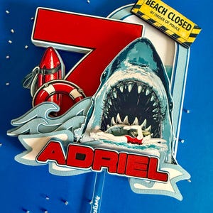 May include: A layered cake topper featuring a large red number 7, a shark with an open mouth, a swimmer in a red swimsuit, a red life buoy, and a yellow sign that reads BEACH CLOSED BY ORDER OF POLICE. The name ADRIEL is printed in red letters.