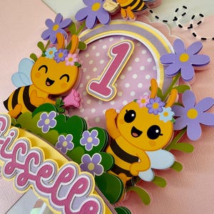 May include: A colorful cake topper featuring cartoon bees, flowers, and the number 1. The topper includes the name "Giselle" in a layered design. The bees are yellow and black with blue wings, and the flowers are purple.