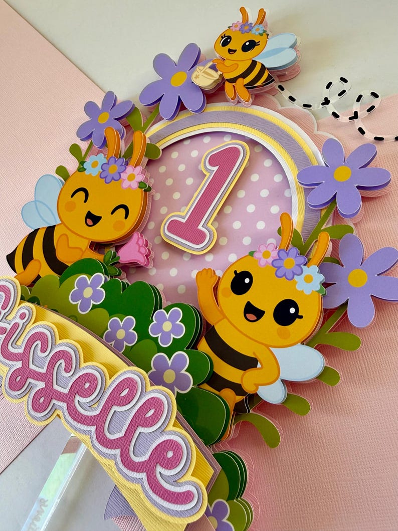 May include: A colorful cake topper featuring cartoon bees, flowers, and the number 1. The topper includes the name "Giselle" in a layered design. The bees are yellow and black with blue wings, and the flowers are purple.