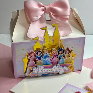 May include: A white favor box with a pink bow and a gold castle cutout. The castle has a group of Disney princesses on it. The box says "Alexa and Angie" on the front.