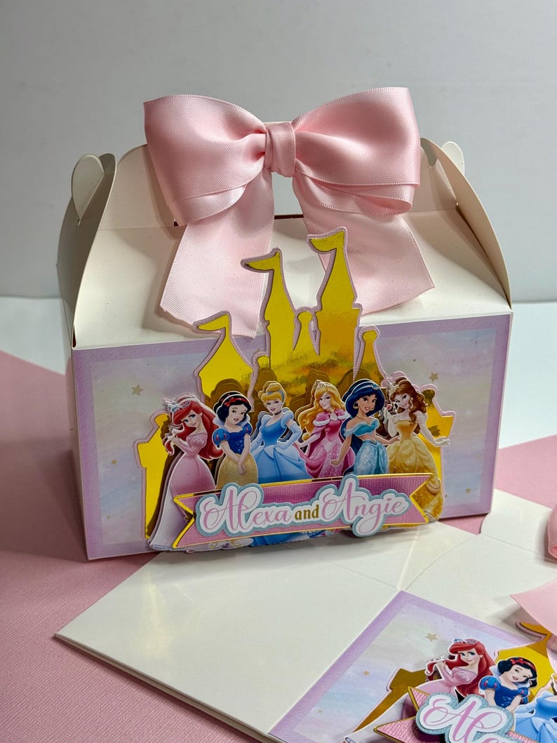 May include: A white favor box with a pink bow and a gold castle cutout. The castle has a group of Disney princesses on it. The box says "Alexa and Angie" on the front.