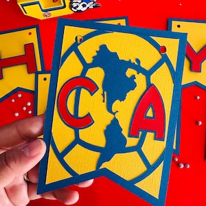 May include: A blue and yellow paper banner with the letters "C" and "A" in red. The banner features a soccer ball design with a blue outline.
