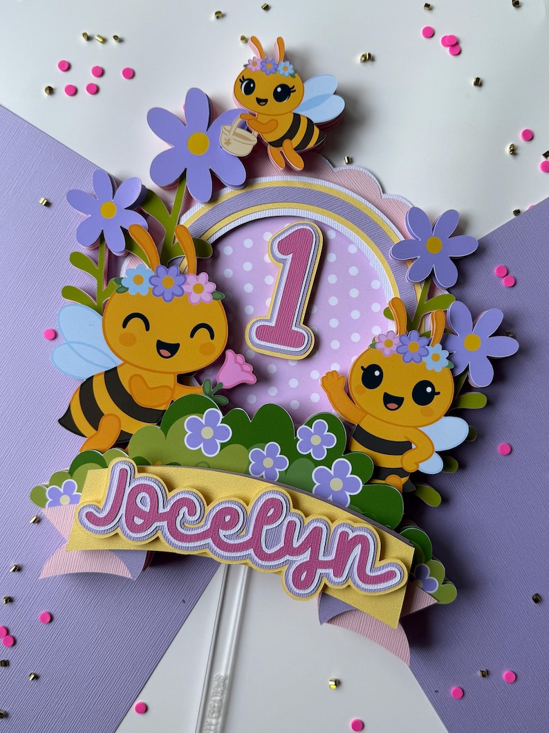 May include: A colorful cake topper featuring cartoon bees, flowers, and the name "Jocelyn". The number "1" is prominently displayed, suggesting a first birthday celebration. The design includes a yellow banner and a clear stick for insertion.