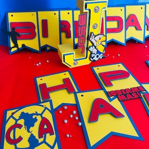 May include: A yellow and blue birthday banner with the words "Birthday" and "Happy" in red letters. The banner also features a large letter "J" with the words "&iexcl;odiame mas!" and a cartoon soccer player.
