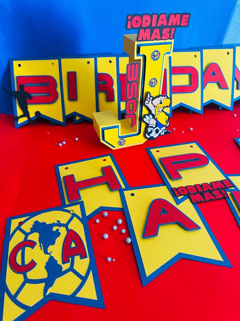 May include: A yellow and blue birthday banner with the words "Birthday" and "Happy" in red letters. The banner also features a large letter "J" with the words "&iexcl;odiame mas!" and a cartoon soccer player.