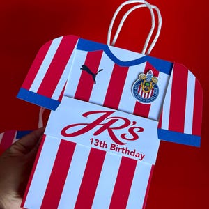May include: A gift bag shaped like a soccer jersey, featuring red and white stripes with blue accents. The bag has a Puma logo and a team emblem. The text "JR's 13th Birthday" is displayed on the front.