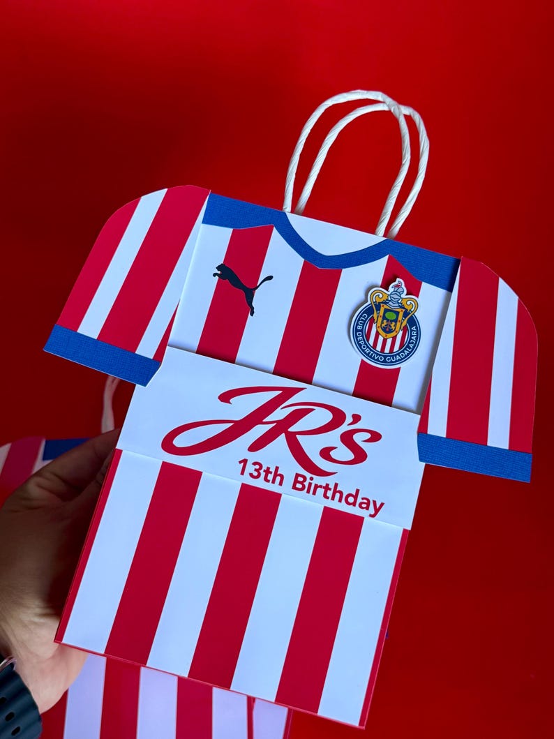 May include: A gift bag shaped like a soccer jersey, featuring red and white stripes with blue accents. The bag has a Puma logo and a team emblem. The text "JR's 13th Birthday" is displayed on the front.