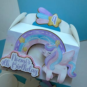 May include: A white party favor box decorated with a unicorn, rainbow, and shooting star design. The box features the text "Noemi's 4th Birthday" in a cursive font. The unicorn has a pink and purple mane and tail.