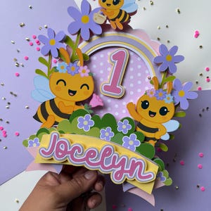 May include: A colorful cake topper featuring cartoon bees, flowers, and the number 1. The topper includes the name "Jocelyn" in pink script. The design is layered with various colors, including yellow, purple, and green, creating a festive look.