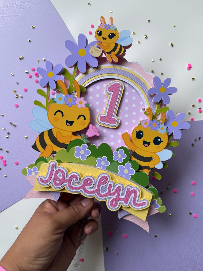 May include: A colorful cake topper featuring cartoon bees, flowers, and the number 1. The topper includes the name "Jocelyn" in pink script. The design is layered with various colors, including yellow, purple, and green, creating a festive look.