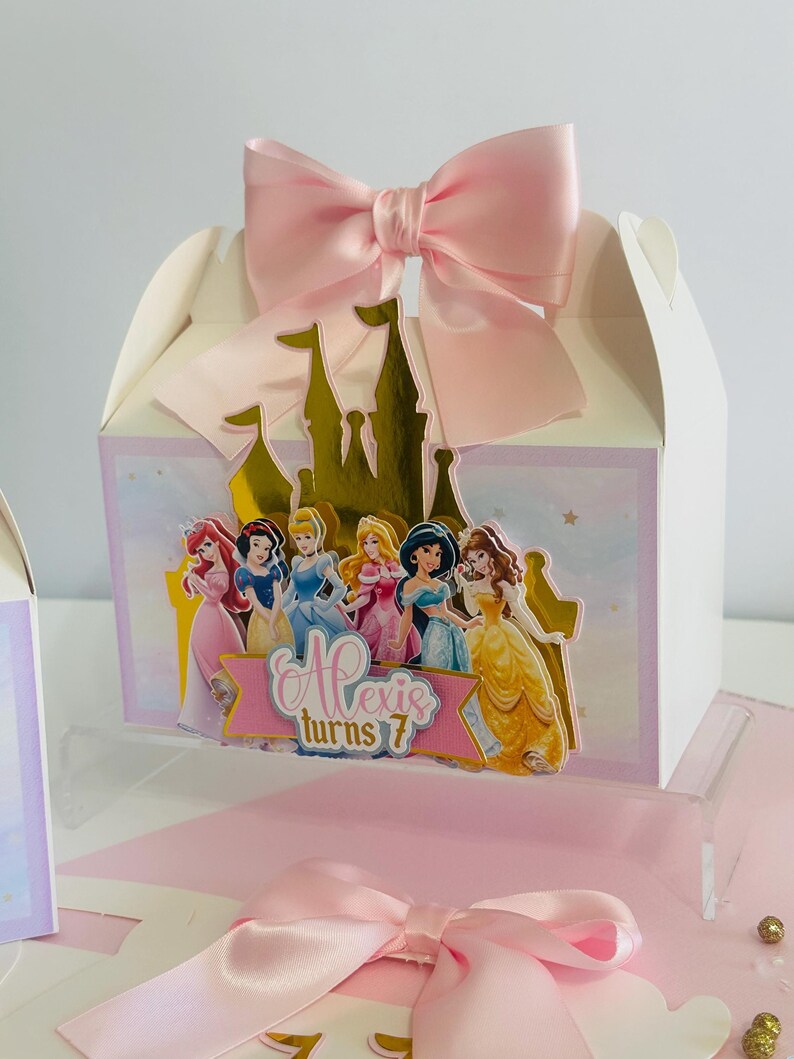 May include: A white favor box with a pink bow and a gold castle design. The box features a picture of Disney princesses and the text "Alexis turns 7".