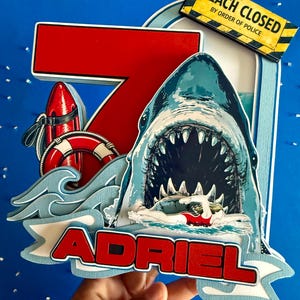 May include: A layered cake topper featuring a large red number seven, a shark with an open mouth, a swimmer, a red life buoy, and a red rescue float. A yellow sign reads BEACH CLOSED BY ORDER OF POLICE. A banner at the bottom displays the name ADRIEL.