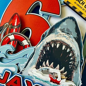 May include: A layered paper cake topper featuring a large red number 6, a shark with an open mouth, a swimmer, a red life buoy, and a red rescue tube. The name JAXON is printed in red and white letters. A yellow sign reads BEACH CLOSED BY ORDER OF POLICE.