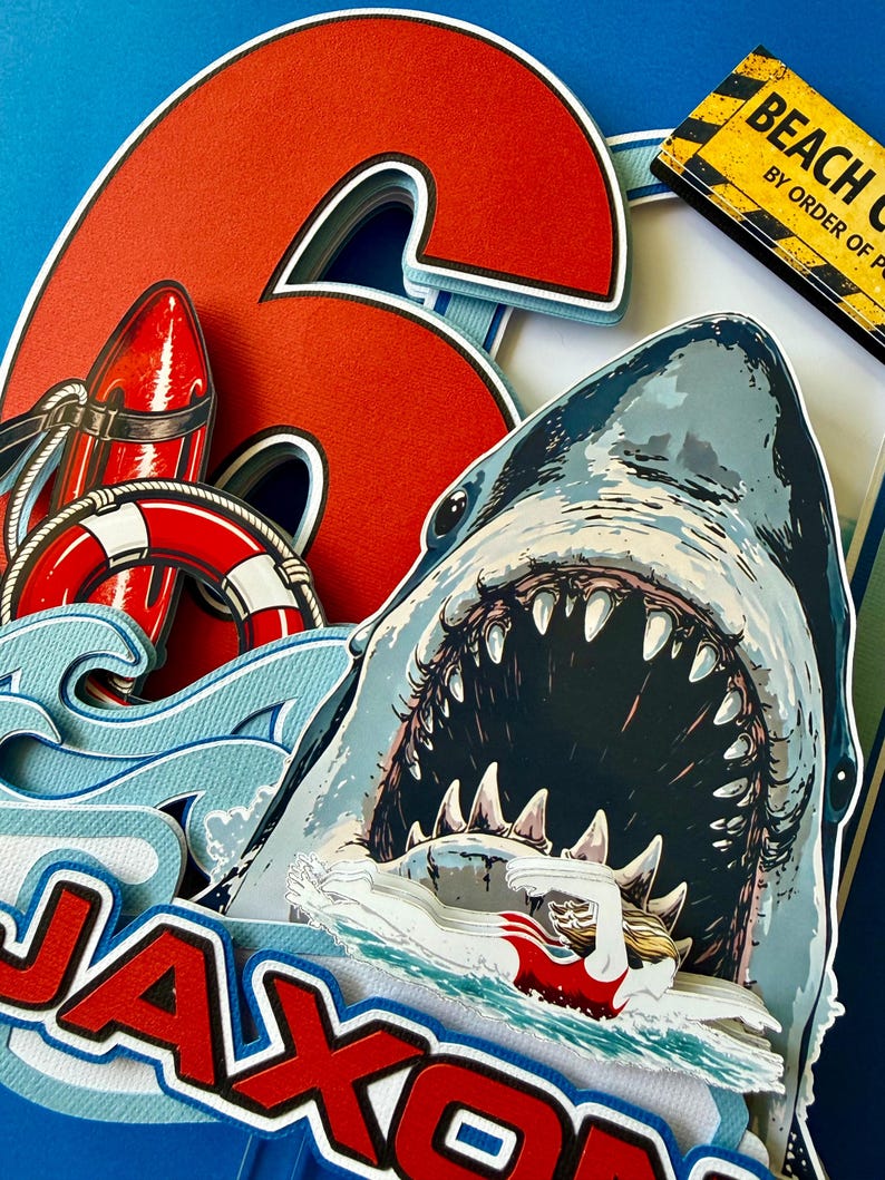 May include: A layered paper cake topper featuring a large red number 6, a shark with an open mouth, a swimmer, a red life buoy, and a red rescue tube. The name JAXON is printed in red and white letters. A yellow sign reads BEACH CLOSED BY ORDER OF POLICE.