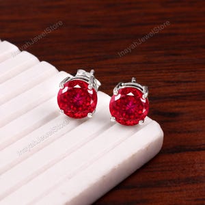 May include: A pair of round-cut, vibrant red gemstone stud earrings set in polished silver-toned metal prongs. Each earring features a faceted circular stone that reflects light, secured by a standard post backing for pierced ears.