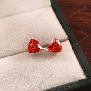 Puede incluir: Un par de pendientes de botón con gemas triangulares de color naranja brillante engastadas en plata. Las gemas tienen un aspecto facetado. Los pendientes se exhiben en una caja de joyería gris.
