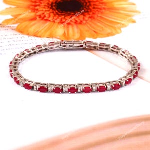 May include: A silver bracelet with a line of oval-cut, bright red gemstones, with small, clear stones between them. The bracelet is shown on a white surface, with a blurred orange flower in the background. The jewelry is from InayahJewelStore.