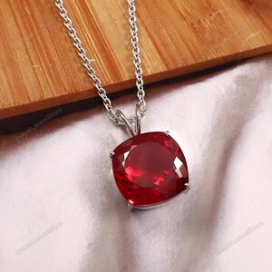 May include: A silver pendant necklace featuring a large, square-cut, deep red gemstone. The gemstone is set in a silver bezel and hangs from a delicate silver chain. The necklace is displayed on a white surface with a wooden background.