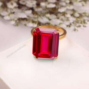 May include: A gold-toned ring featuring a large, rectangular, emerald-cut vibrant red gemstone set in a four-prong setting. The band is smooth and metallic, providing a clean contrast to the faceted, deep red stone that dominates the design.