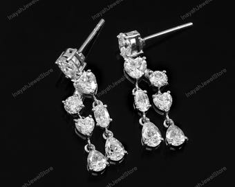 May include: A pair of silver dangle earrings with multiple clear, sparkling gemstones. The earrings feature a stud at the top with a large, round stone, followed by a series of heart, oval, and pear-shaped stones cascading downwards.