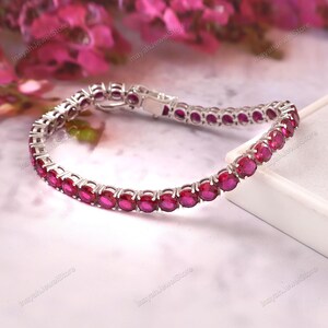 May include: A silver bracelet featuring a series of oval-cut, vibrant red gemstones. The bracelet is set in a delicate, linked design with a secure clasp. The gemstones are evenly spaced, creating a continuous line of color. The background includes pink flowers.