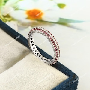 May include: A silver ring with a continuous band of small, round, red gemstones and clear gemstones. The ring is set in a silver band with a geometric pattern on the sides. The ring is displayed in a blue jewelry box.