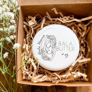 Op de afbeelding: Een witte pot Bala Butter, 113g, in een kartonnen doos met papieren vulling. De pot heeft een zwart bloemmotief en de woorden "Bala Mama" en "Bala Butter" erop. Witte bloemen op de achtergrond.
